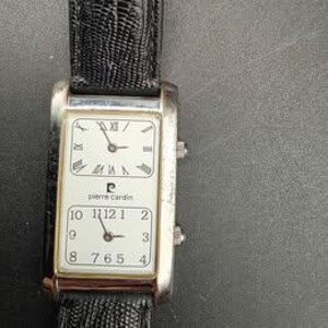 Puede incluir: Un reloj de pulsera rectangular Pierre Cardin con caja plateada y correa de cuero negro. La esfera del reloj est&aacute; dividida en dos secciones, una con n&uacute;meros romanos y otra con n&uacute;meros ar&aacute;bigos. El reloj muestra la hora.