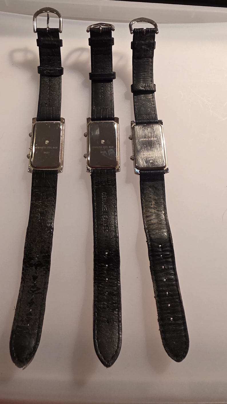 Puede incluir: Tres relojes de pulsera con correas de cuero negro y esferas rectangulares plateadas. Las correas de los relojes son texturizadas y tienen hebillas plateadas. Las esferas de los relojes tienen la marca "Pierre Cardin" y "Stainless Steel Back".