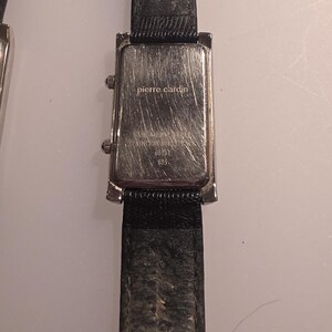Puede incluir: Primer plano de una correa de reloj de cuero negro con una esfera rectangular plateada. La esfera del reloj tiene la inscripci&oacute;n "pierre cardin". El reloj es de estilo cl&aacute;sico.