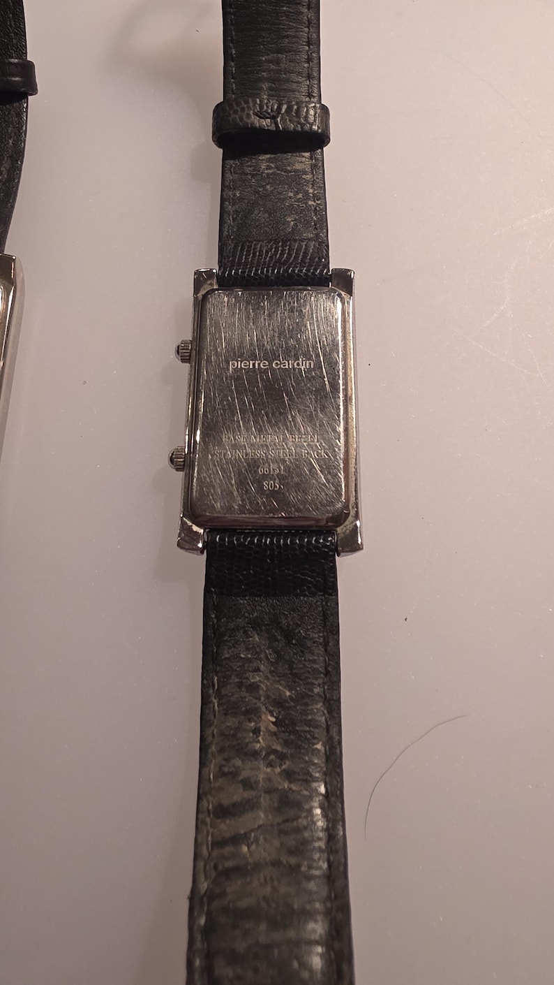 Puede incluir: Primer plano de una correa de reloj de cuero negro con una esfera rectangular plateada. La esfera del reloj tiene la inscripci&oacute;n "pierre cardin". El reloj es de estilo cl&aacute;sico.