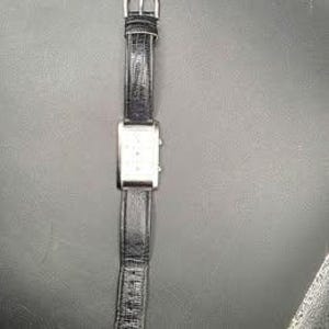 Puede incluir: Un reloj de pulsera rectangular plateado con una correa de cuero negro. La esfera del reloj tiene peque&ntilde;as marcas espaciadas uniformemente. La correa mide aproximadamente 2 cm de ancho. El reloj se muestra sobre un fondo oscuro.