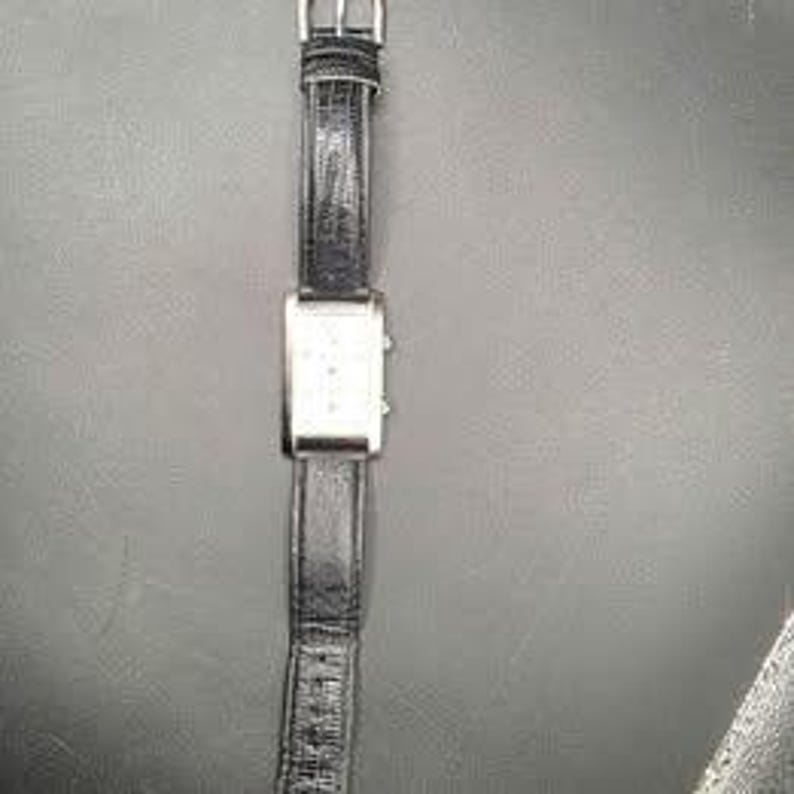 Puede incluir: Un reloj de pulsera rectangular plateado con una correa de cuero negro. La esfera del reloj tiene peque&ntilde;as marcas espaciadas uniformemente. La correa mide aproximadamente 2 cm de ancho. El reloj se muestra sobre un fondo oscuro.