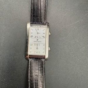 Puede incluir: Un reloj de pulsera rectangular Pierre Cardin con caja plateada y correa de cuero negro. La esfera es blanca con n&uacute;meros romanos en la parte superior y n&uacute;meros ar&aacute;bigos en la inferior. La correa mide aproximadamente 2 cm de ancho.