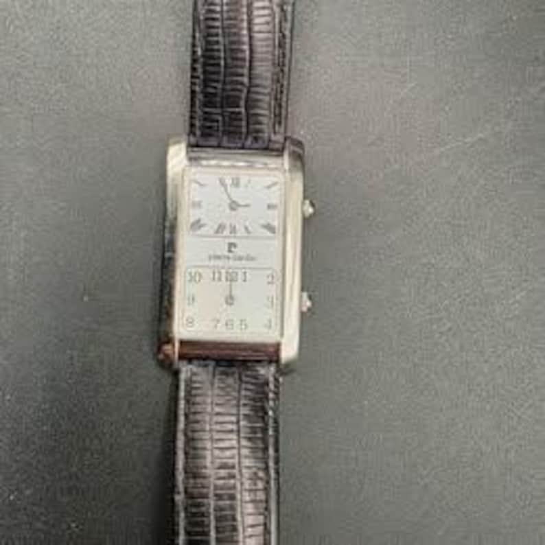 Puede incluir: Un reloj de pulsera rectangular Pierre Cardin con caja plateada y correa de cuero negro. La esfera es blanca con n&uacute;meros romanos en la parte superior y n&uacute;meros ar&aacute;bigos en la inferior. La correa mide aproximadamente 2 cm de ancho.