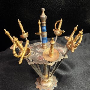 May include: A metal hors d'oeuvre holder featuring a central pillar with a blue ringed accent, supporting a square platform with decorative floral patterns. Six gold-toned sword-shaped picks are inserted into the platform, each with a detailed hilt.