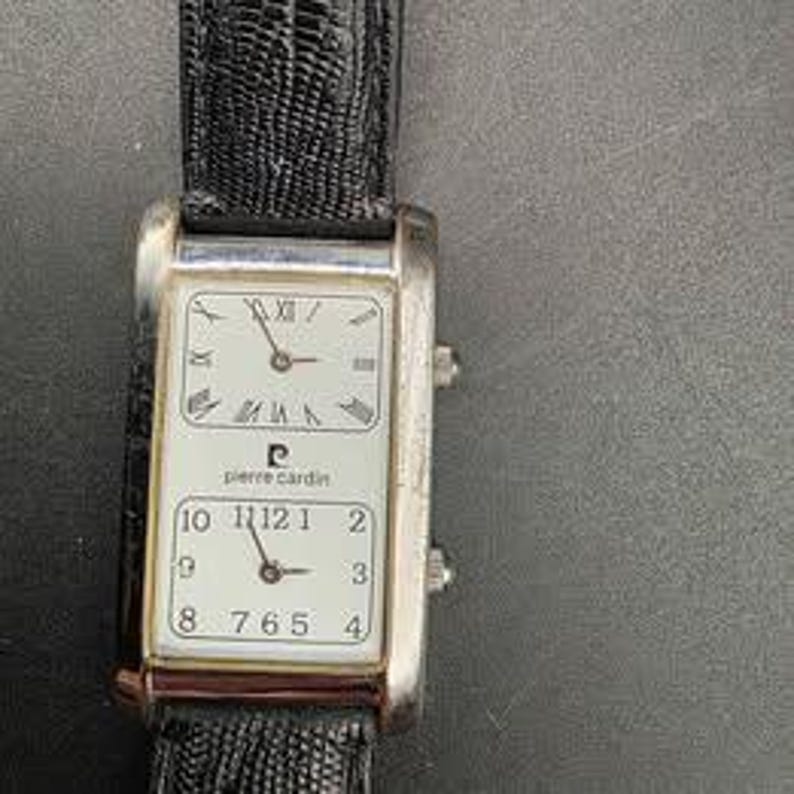 Puede incluir: Un reloj de pulsera Pierre Cardin rectangular con caja plateada y correa de cuero negro. La esfera del reloj tiene dos esferas separadas, una con n&uacute;meros romanos y otra con n&uacute;meros ar&aacute;bigos. El reloj muestra la hora.