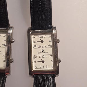 Puede incluir: Dos relojes Pierre Cardin rectangulares con correas de cuero negro. Las esferas blancas muestran n&uacute;meros romanos y ar&aacute;bigos. El nombre de la marca est&aacute; impreso en la esfera.