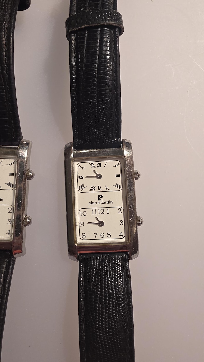 Puede incluir: Dos relojes Pierre Cardin rectangulares con correas de cuero negro. Las esferas blancas muestran n&uacute;meros romanos y ar&aacute;bigos. El nombre de la marca est&aacute; impreso en la esfera.