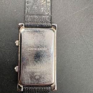 Puede incluir: Un reloj rectangular Pierre Cardin con caja plateada y correa de cuero negro. La esfera del reloj no es visible. La parte trasera del reloj tiene el logotipo de Pierre Cardin y otro texto.