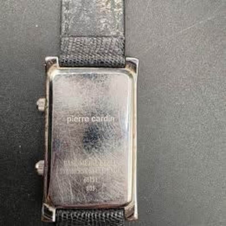 Puede incluir: Un reloj rectangular Pierre Cardin con caja plateada y correa de cuero negro. La esfera del reloj no es visible. La parte trasera del reloj tiene el logotipo de Pierre Cardin y otro texto.