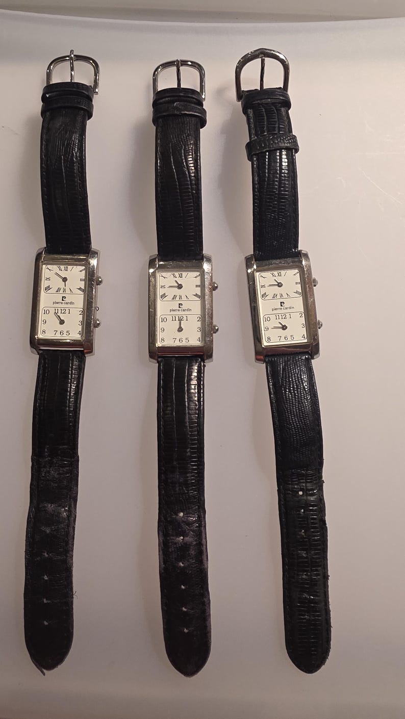 Puede incluir: Tres relojes de pulsera rectangulares con correas de cuero negro. Las esferas son blancas con n&uacute;meros y manecillas negras, y la marca "pierre cardin" est&aacute; impresa en la esfera. Las correas tienen hebillas plateadas.