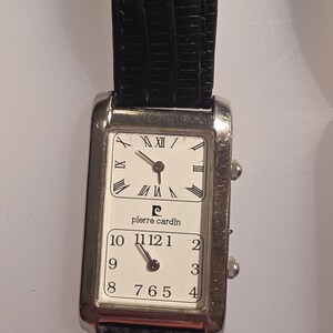 Puede incluir: Reloj de pulsera Pierre Cardin rectangular con correa de cuero negro. La esfera del reloj est&aacute; dividida en dos secciones, cada una con fondo blanco y n&uacute;meros romanos negros. El reloj tiene dos peque&ntilde;os diales en el lateral.