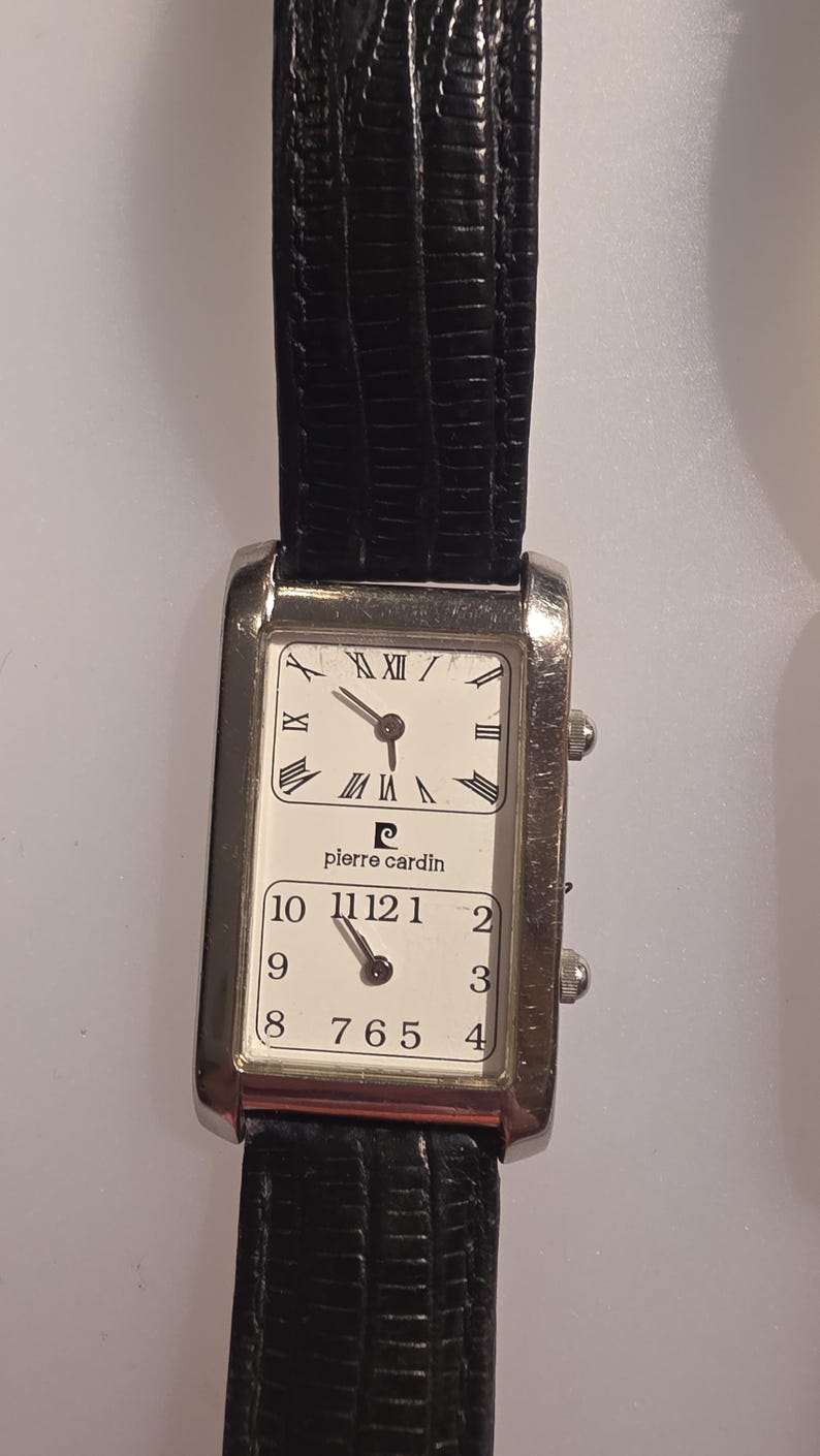 Puede incluir: Reloj de pulsera Pierre Cardin rectangular con correa de cuero negro. La esfera del reloj est&aacute; dividida en dos secciones, cada una con fondo blanco y n&uacute;meros romanos negros. El reloj tiene dos peque&ntilde;os diales en el lateral.