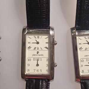 Puede incluir: Reloj Pierre Cardin rectangular con correa de cuero negro. La esfera del reloj est&aacute; dividida en dos secciones, con n&uacute;meros romanos en la parte superior y ar&aacute;bigos en la inferior. La caja del reloj es plateada.
