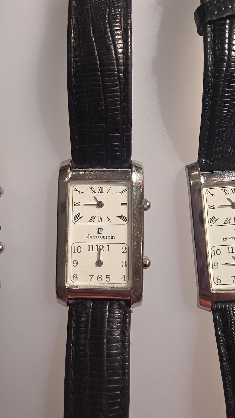 Puede incluir: Reloj Pierre Cardin rectangular con correa de cuero negro. La esfera del reloj est&aacute; dividida en dos secciones, con n&uacute;meros romanos en la parte superior y ar&aacute;bigos en la inferior. La caja del reloj es plateada.
