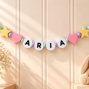 May include: A decorative wall hanging garland featuring wooden beads and shapes strung on a rope. The garland includes blue clouds, yellow stars, pink hearts, and white circular beads with the black letters A, R, I, A printed on them.
