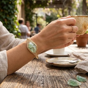 May include: A gold wire-wrapped cuff bracelet featuring a central raw, light green fluorite crystal. The bracelet is worn on a wrist, and a thin gold ring is visible on the finger of the same hand, which is holding a speckled ceramic mug.