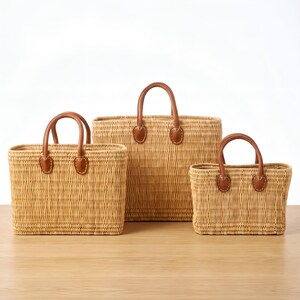 May include: Three rectangular woven baskets of varying sizes, each crafted from light tan natural fibers. Each basket features two sturdy brown leather handles securely attached with visible stitching to the front and back panels.