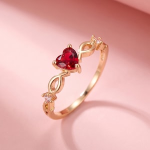 May include: A gold-colored ring featuring a heart-shaped, deep red gemstone as the focal point. The ring has a delicate design with small, clear accent stones and an intricate band. The ring is set against a soft pink background.