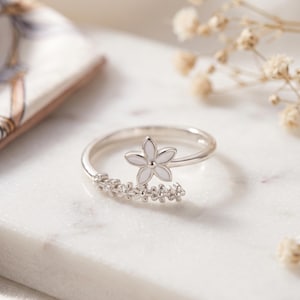 Peut inclure: Une bague argentée avec un motif de fleur blanche. La bague présente une grande fleur en haut et une rangée de petites fleurs autour du bracelet. La bague est posée sur une surface en marbre blanc.