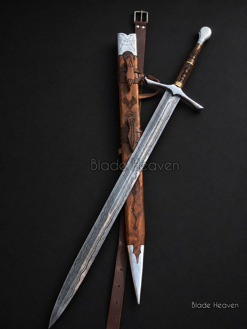 May include: A double-edged steel sword with a wavy pattern along the blade, a silver crossguard, and a brown wooden handle. It is accompanied by a matching brown wooden scabbard featuring silver metal accents and a brown leather belt strap.