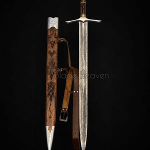 May include: A longsword with a patterned steel blade, a dark wood grip, and a silver pommel. Beside it is a matching wooden scabbard featuring intricate engravings, silver metal fittings, and a brown leather strap with a metal buckle. Blade Heaven.