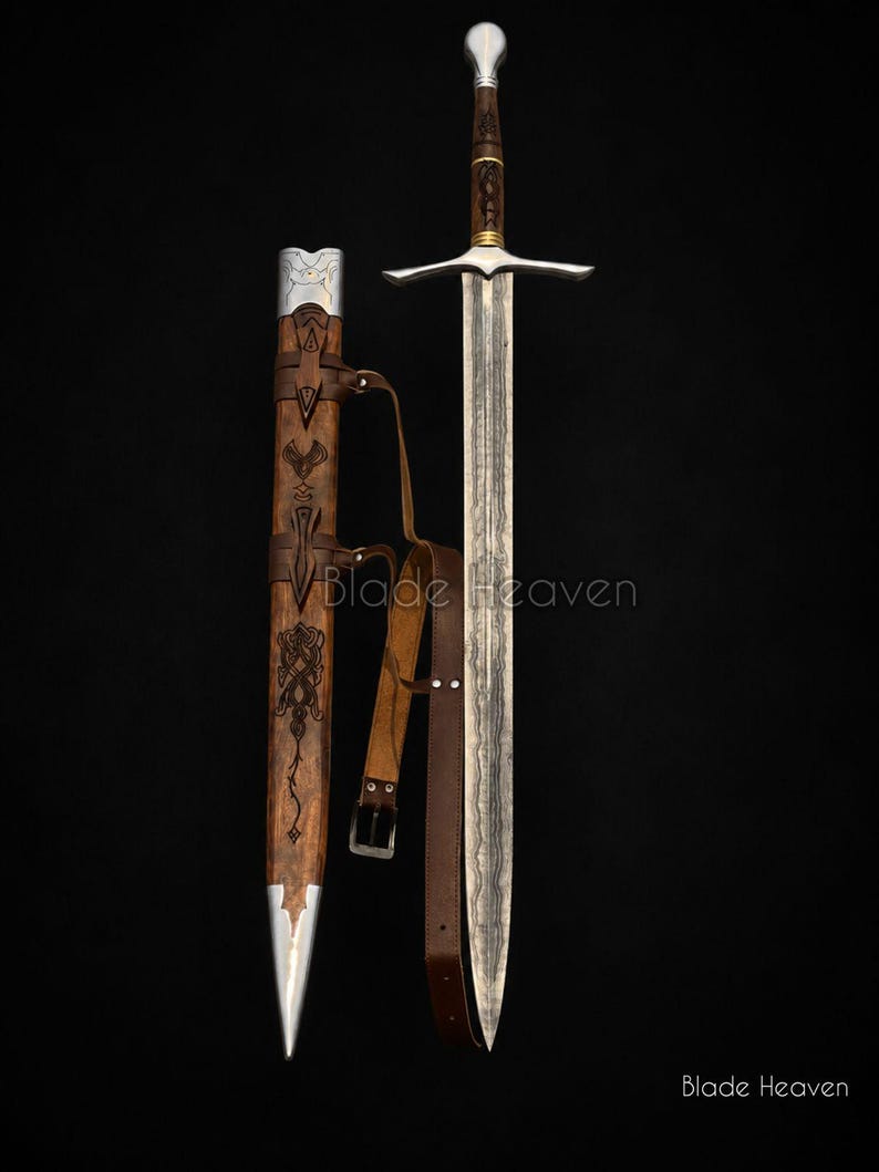 May include: A longsword with a patterned steel blade, a dark wood grip, and a silver pommel. Beside it is a matching wooden scabbard featuring intricate engravings, silver metal fittings, and a brown leather strap with a metal buckle. Blade Heaven.