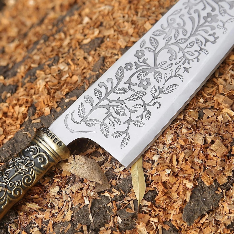 May include: A kitchen knife with a silver-toned blade featuring an etched floral and vine pattern. The handle is made of dark, textured metal with an ornate embossed design and the word MASTER stamped clearly on the bolster near the blade.