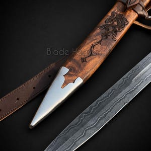 May include: A dagger with a patterned Damascus steel blade and a wooden scabbard featuring intricate carvings and a polished metal tip. A brown leather strap is attached to the scabbard. The text Blade Heaven is visible on the image.