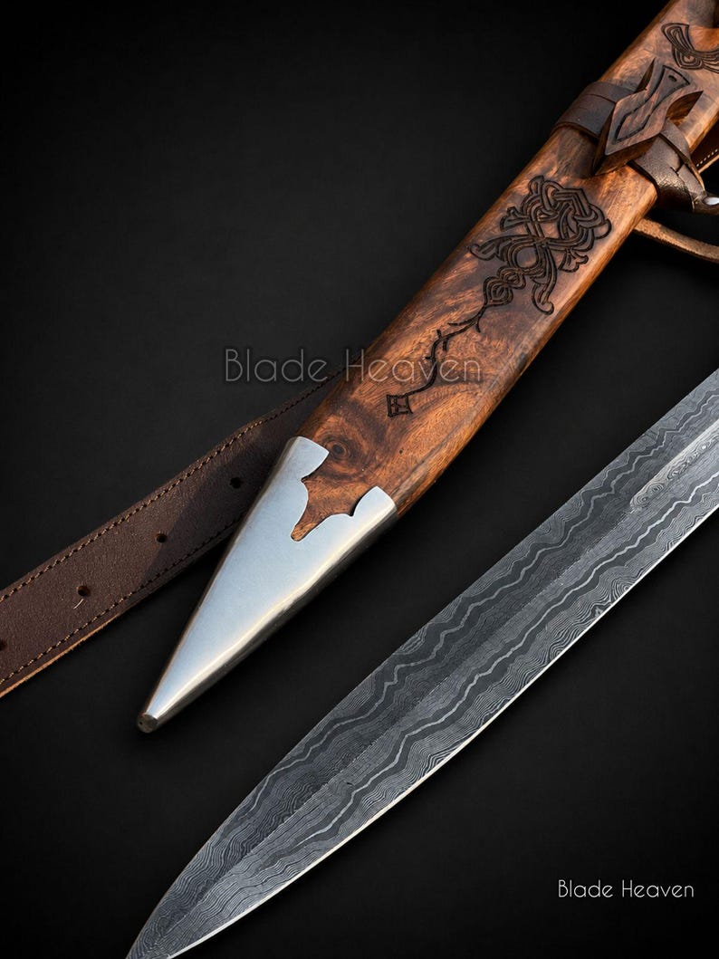 May include: A dagger with a patterned Damascus steel blade and a wooden scabbard featuring intricate carvings and a polished metal tip. A brown leather strap is attached to the scabbard. The text Blade Heaven is visible on the image.
