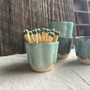 Pode incluir: Vários porta-fósforos de cerâmica em tons de azul e verde, um deles cheio de fósforos. Os recipientes têm um design ondulado e uma base creme. Fundo de madeira. Medidas em cm.