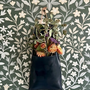 May include: A dark blue leather wall pocket filled with dried flowers in shades of orange, purple, and green. The pocket hangs against a dark green floral wallpaper background.
