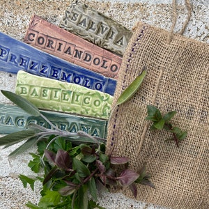 May include: Set of six ceramic herb markers with Italian names for herbs: Salvia, Coriandolo, Prezzemolo, Basilico, Maggiorana. The markers are arranged in a stack with a burlap bag and fresh herbs in the background.