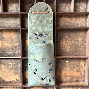 May include: A tall, semi-circular ceramic wall pocket with a rounded top and a leather hanging cord. The surface features a mottled glaze in shades of sage green, cream, and dark blue, creating a textured, speckled appearance across the entire piece.