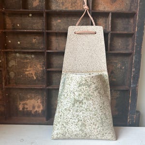 Puede incluir: Un bolsillo de pared de cerámica hecho a mano con un diseño moteado de dos tonos. La parte superior es de color beige claro, mientras que la inferior es verde moteado. Se adjunta un cordón de cuero para colgar. Ideal para plantas pequeñas.