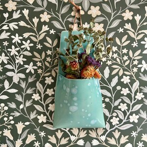 Op de afbeelding: Een turquoise keramische wandzak met een leren riem, gevuld met gedroogde bloemen in tinten oranje, paars en groen. De zak hangt tegen een donkergroen bloemenbehang, wat een rustieke en decoratieve uitstraling creëert.