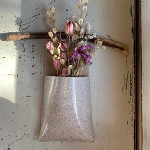 Puede incluir: Un pequeño jarrón de cerámica blanca moteado con un arreglo de flores secas. Las flores son una mezcla de rosa, morado y marrón.
