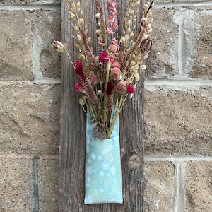 May include: A wall-mounted display featuring a vertical weathered wooden board holding a light blue ceramic pocket vase. The vase contains a dried floral arrangement with pink, magenta, and tan stems, including seed pods and various textured grasses.