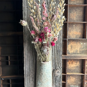 Puede incluir: Una pared de madera rústica con un jarrón de cerámica fijado a ella. El jarrón está lleno de flores secas en tonos de rosa, rojo y beige.