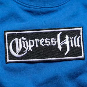 May include: A bright blue sweatshirt featuring a rectangular black patch centred on the chest. The patch has a white embroidered border and displays the text Cypress Hill in a stylised white gothic font across the black fabric background.