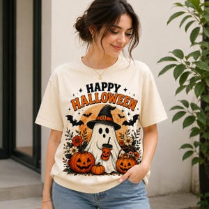 May include: A cream-colored short-sleeve t-shirt featuring a graphic print with the text "HAPPY HALLOWEEN". The design includes a ghost wearing a witch hat, holding a coffee cup, surrounded by pumpkins, flowers, bats, and a full moon.