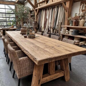 May include: A substantial, reclaimed wooden dining table with a natural finish, accompanied by woven wicker chairs. The table features a thick, live-edge top and robust wooden legs. The setting is in a bright, open space with various decorative items.