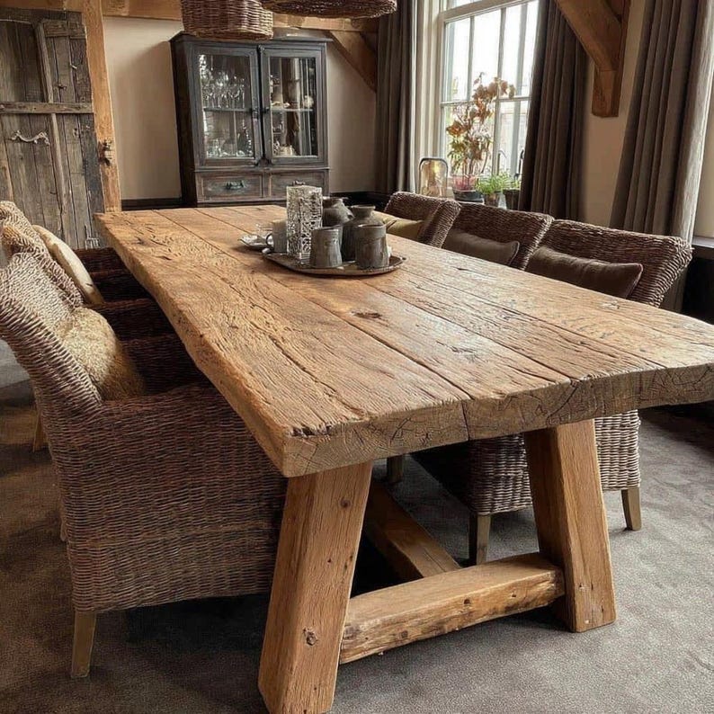 May include: A large, rustic wooden dining table with a natural, weathered finish. The table is supported by thick, angled wooden legs. Woven wicker chairs surround the table, creating a warm dining area. Decorative items sit on the table.