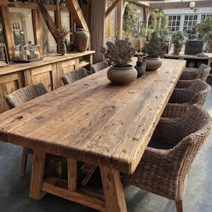 May include: A large, rectangular wooden dining table with a natural, weathered finish. The table is supported by sturdy, angled legs and surrounded by woven wicker chairs. Decorative pots with plants sit on the table. The table measures approximately 305 cm long.