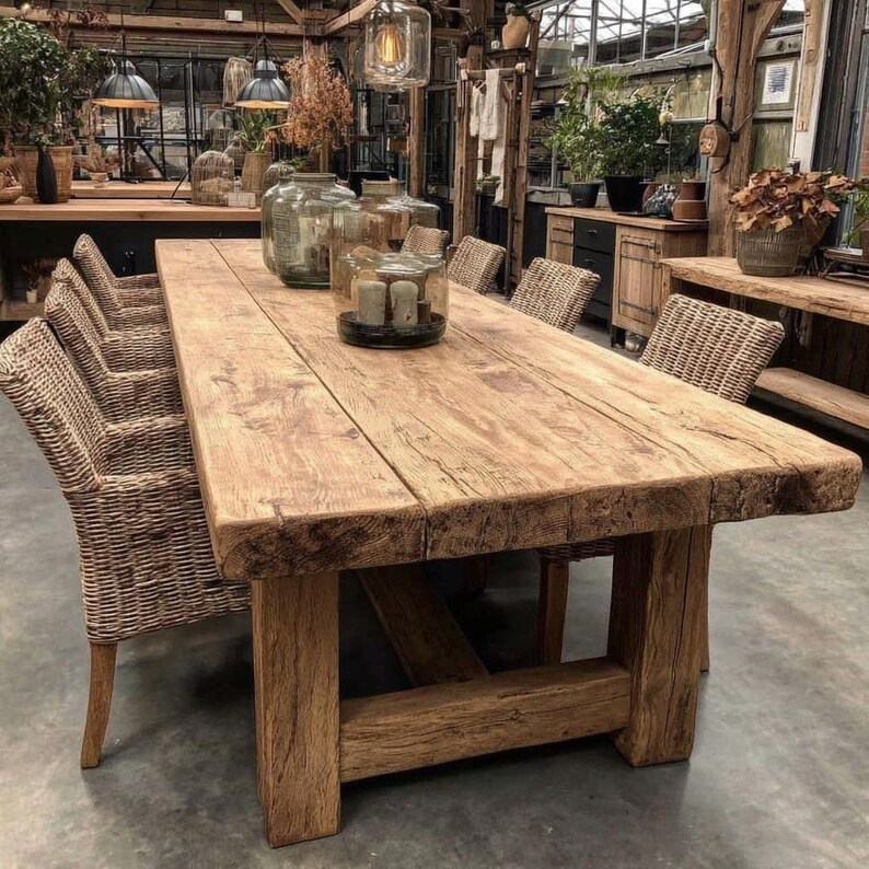 May include: A substantial, rectangular wooden dining table with a distressed, aged look. The table is supported by thick, square legs and surrounded by woven wicker chairs. Decorative glass jars and candles are placed on the table's surface, creating a warm atmosphere.