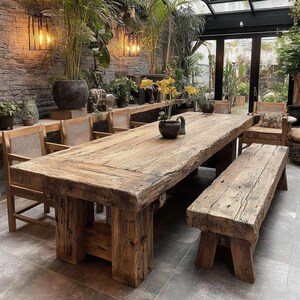 May include: A large, weathered wooden dining table with a matching bench and chairs. The table is made from thick, reclaimed wood and has a natural, unfinished appearance. The chairs have woven backs. The setting is a sunroom with plants and hanging lights.