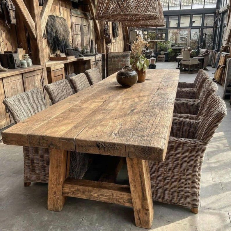 May include: A large, rustic wooden dining table with a weathered appearance. The table is supported by thick, sturdy wooden legs. Wicker chairs surround the table, creating a warm dining area. The table is approximately 3 metres long.