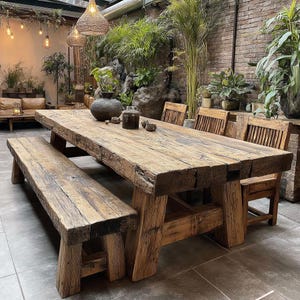 May include: A large, weathered wooden dining table with a matching bench and chairs. The table and bench are constructed from thick, reclaimed wood, displaying natural grain and knots. The chairs have slatted backs. The setting is an outdoor patio with plants and hanging lights.