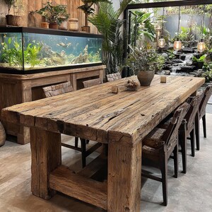 May include: A substantial, reclaimed wooden dining table with a rectangular top and chunky, square legs. Matching wooden chairs surround the table. The room includes a large aquarium, plants, and a waterfall, creating a natural, inviting atmosphere.