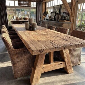May include: A large, rustic wooden dining table with a natural finish. The table has a thick, rectangular top and sturdy A-frame legs. Woven wicker chairs surround the table. The room has large windows and decorative items.